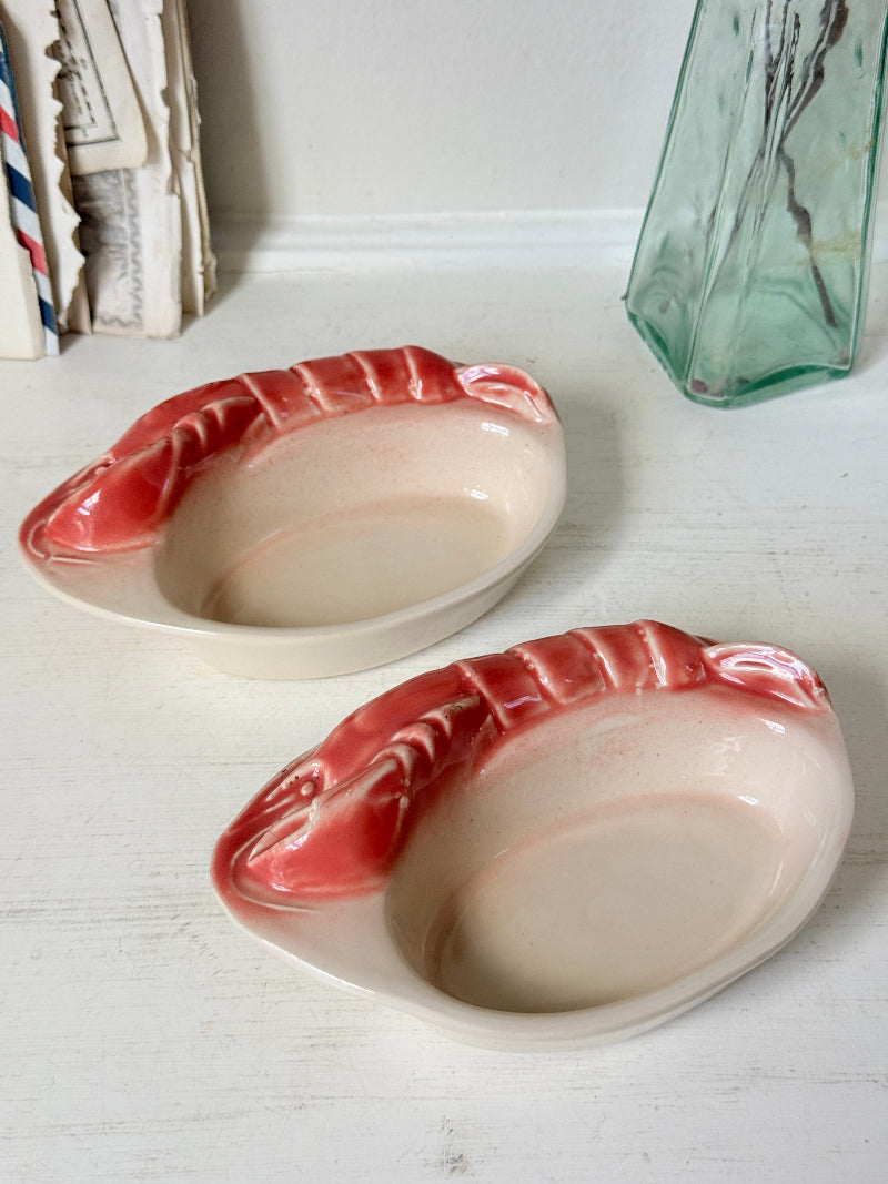 Two ceramic lobster-shaped dish holders with a glossy pink and cream glaze, displayed on a white wooden surface next to a green glass vase.