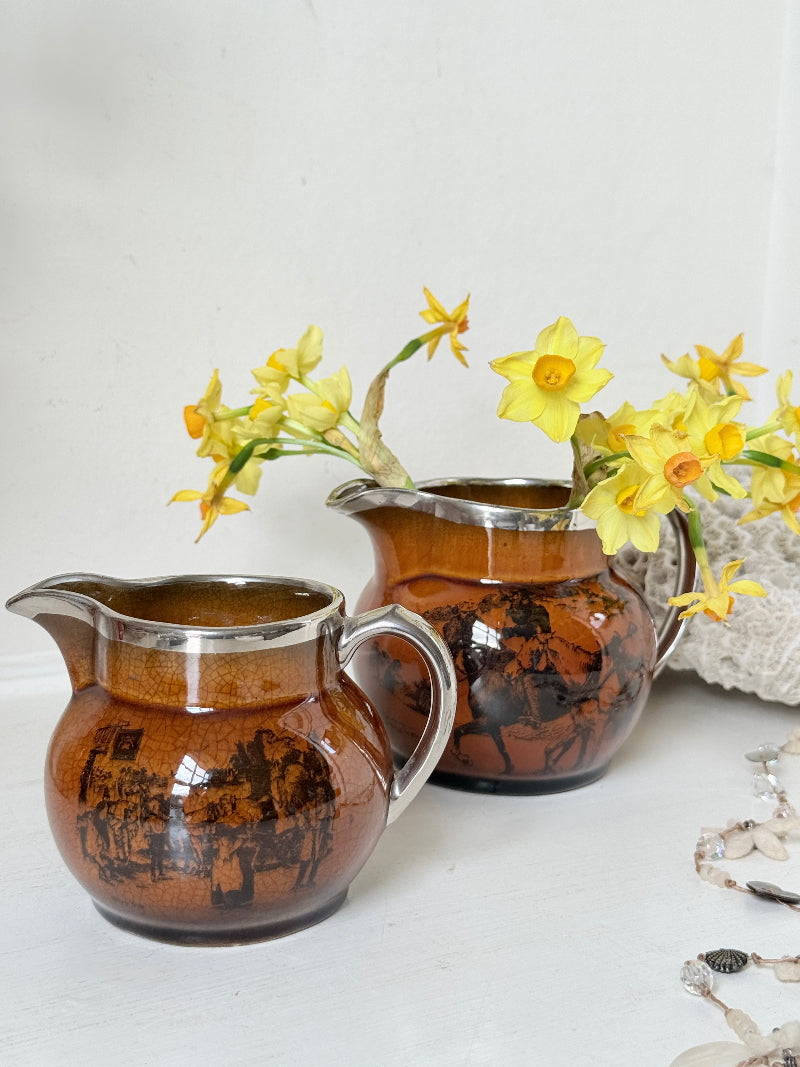 Pair of antique Ridgways stoneware creamer jugs photographed on a white surface in amber treacle glaze with black coaching scenes and silver lustre rims