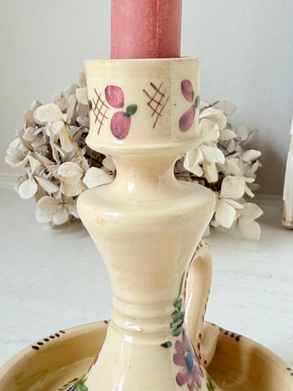 A cream-colored ceramic candlestick holder with painted floral and geometric patterns, holding a pink candle, with dried white flowers in the background.