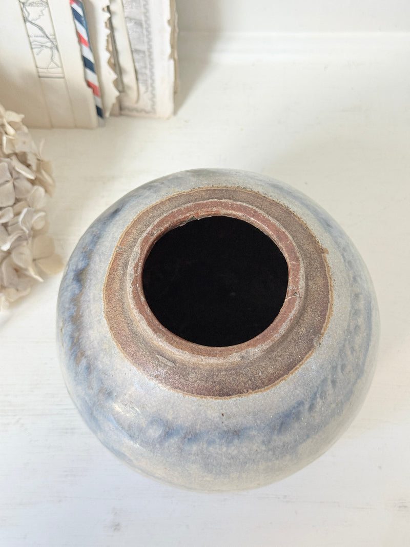 A handmade ceramic pot with a rounded shape, featuring a rustic glaze in shades of blue-gray and brown, photographed on a white wooden surface.
