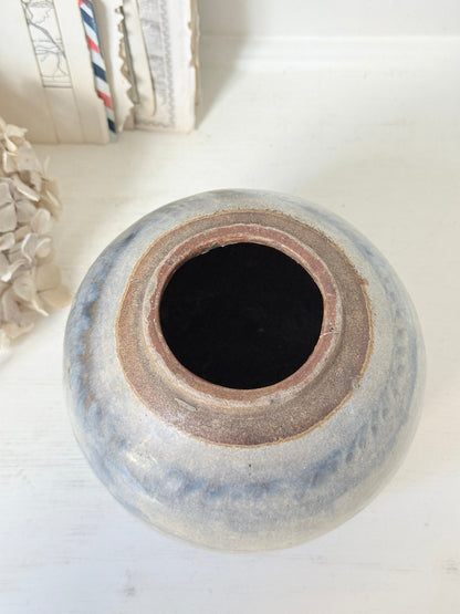 A handmade ceramic pot with a rounded shape, featuring a rustic glaze in shades of blue-gray and brown, photographed on a white wooden surface.