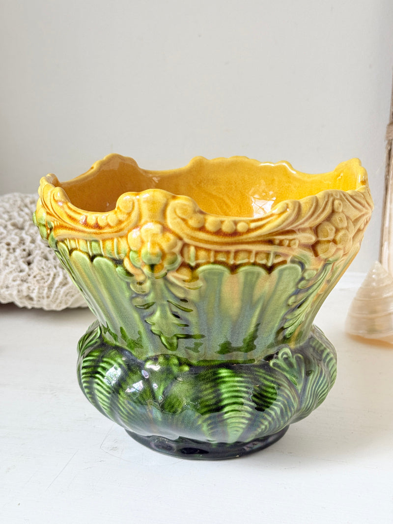 A vintage ceramic planter pot with a yellow and green glazed finish, featuring a decorative leaf and floral pattern, set against a neutral light background.