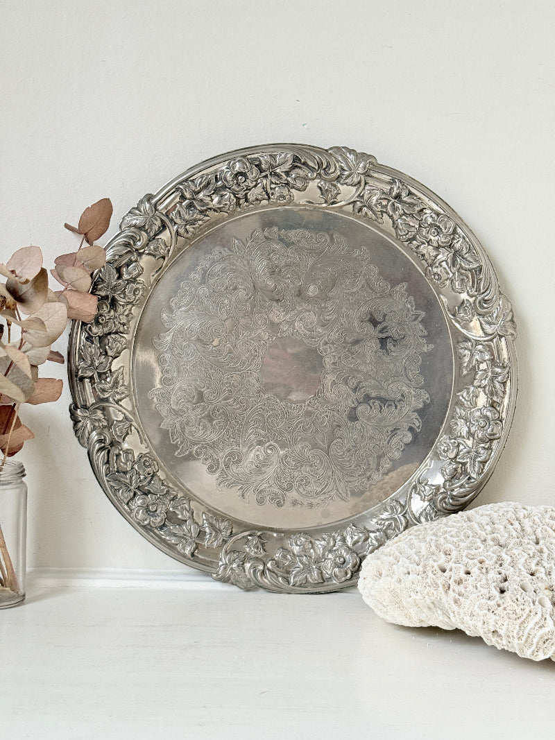 Vintage silver plated round tray with ornate floral border and engraved centre, photographed on a white surface