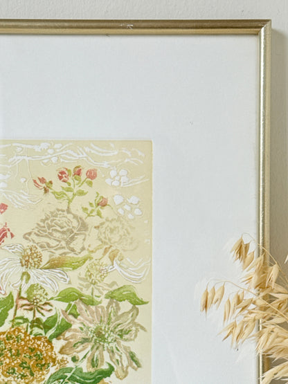 A close-up of a framed botanical art print with a gold frame, featuring cream, green, and pink floral details, styled with dried wheat on a light wall.