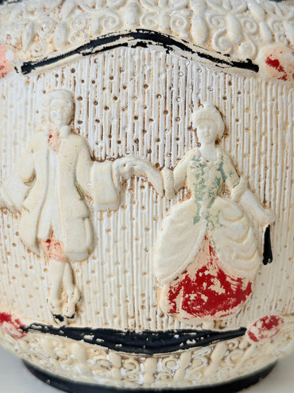Close-up of a vintage ceramic planter featuring embossed figures in a classical style, decorated with red, green, and black accents on an off-white textured background.