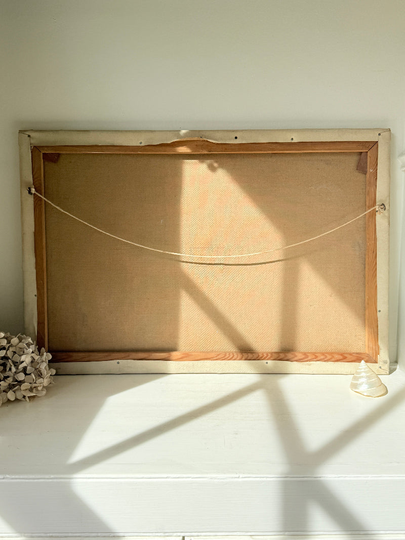 The back of a rectangular framed canvas or print, showing a tan linen ground, wooden frame, and hanging cord, resting on a white shelf with natural window shadows.
