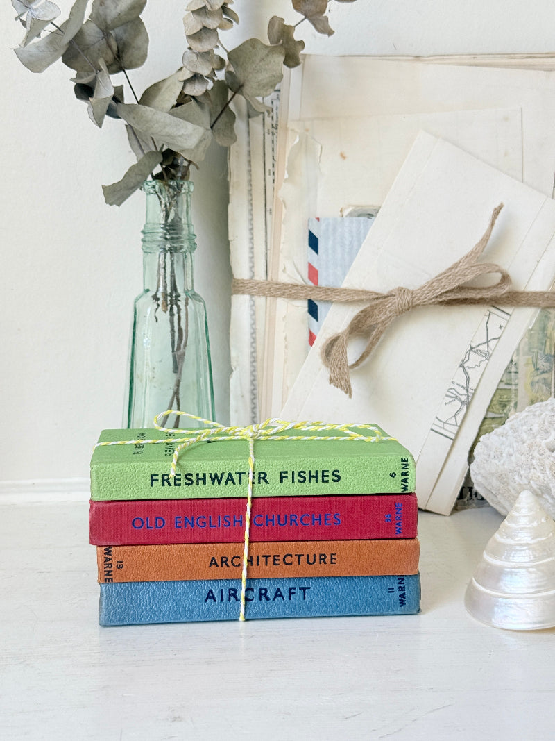 A stack of four colorful hardcover books tied with yellow string, placed on a white surface with a glass bottle vase, dried flowers, and a wrapped book in the background.