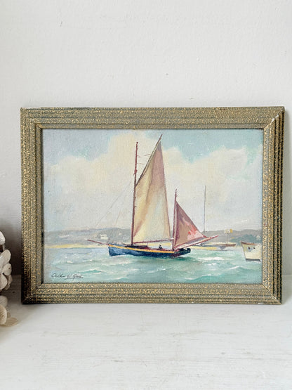 An antique framed painting depicting two sailboats on the water under a cloudy sky, housed in a textured gold frame and displayed on a white surface against a white wall.