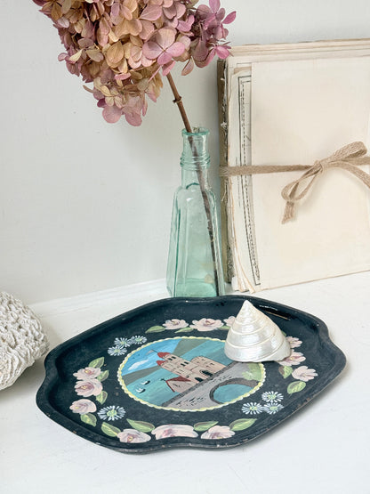 A decorative oval tray with a dark navy blue base, featuring a painted coastal scene in the center and a border of pink flowers and green leaves. A small white seashell sits on the tray.