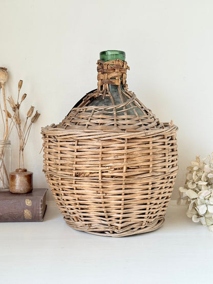 antique French demi john olive oil jar in wicker basket with green glass bottle and painted metal label, displayed on white surface