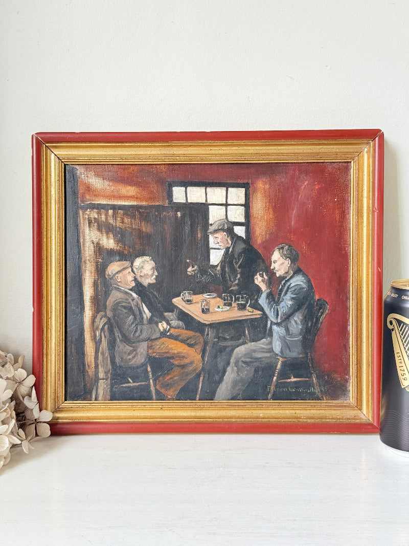 A framed vintage-style oil painting depicting three men sitting around a table in a dimly lit room, with a red and gold frame, displayed on a white surface.