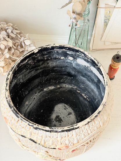 An antique carved ceramic planter pot with a distressed off-white exterior and a dark blackened interior, shown on a white surface with vintage decor in the background.