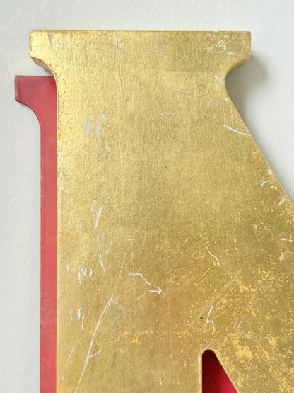 A close-up of a decorative letter cutout featuring a textured gold top surface with visible white scratch-like marks, layered over a red backing against a light neutral background.