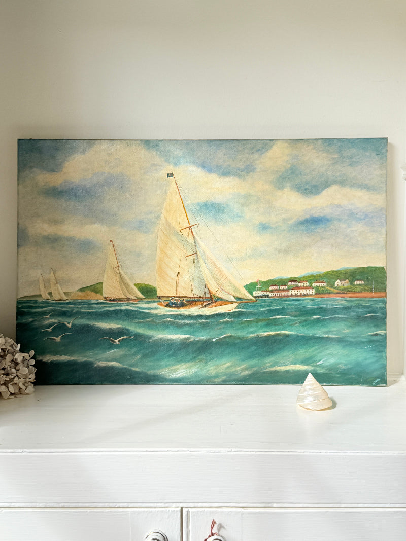 A rectangular oil painting of sailboats on a blue-green sea under a cloudy sky, displayed on a white console table with a small seashell and dried coral decor.