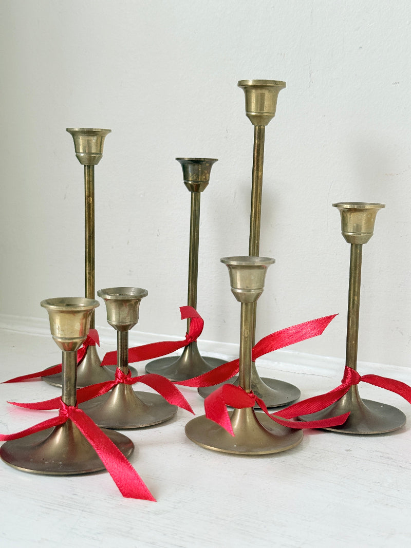 Set of 7 vintage brass candlesticks in graduated heights with aged brass finish styled with red ribbon on white surface