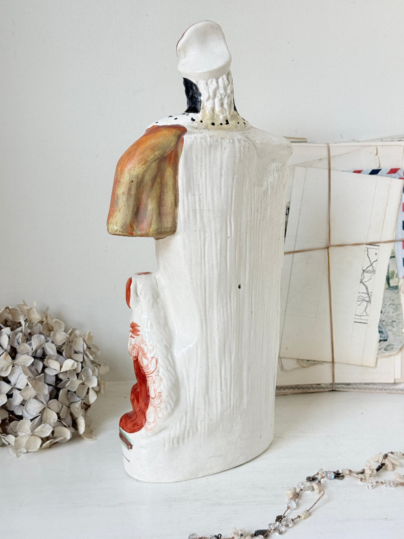 The back of a handmade ceramic figurine with a textured white body, featuring hand-painted abstract details in orange, red, black, and white, styled on a white surface with dried flowers and a string bag in the background.
