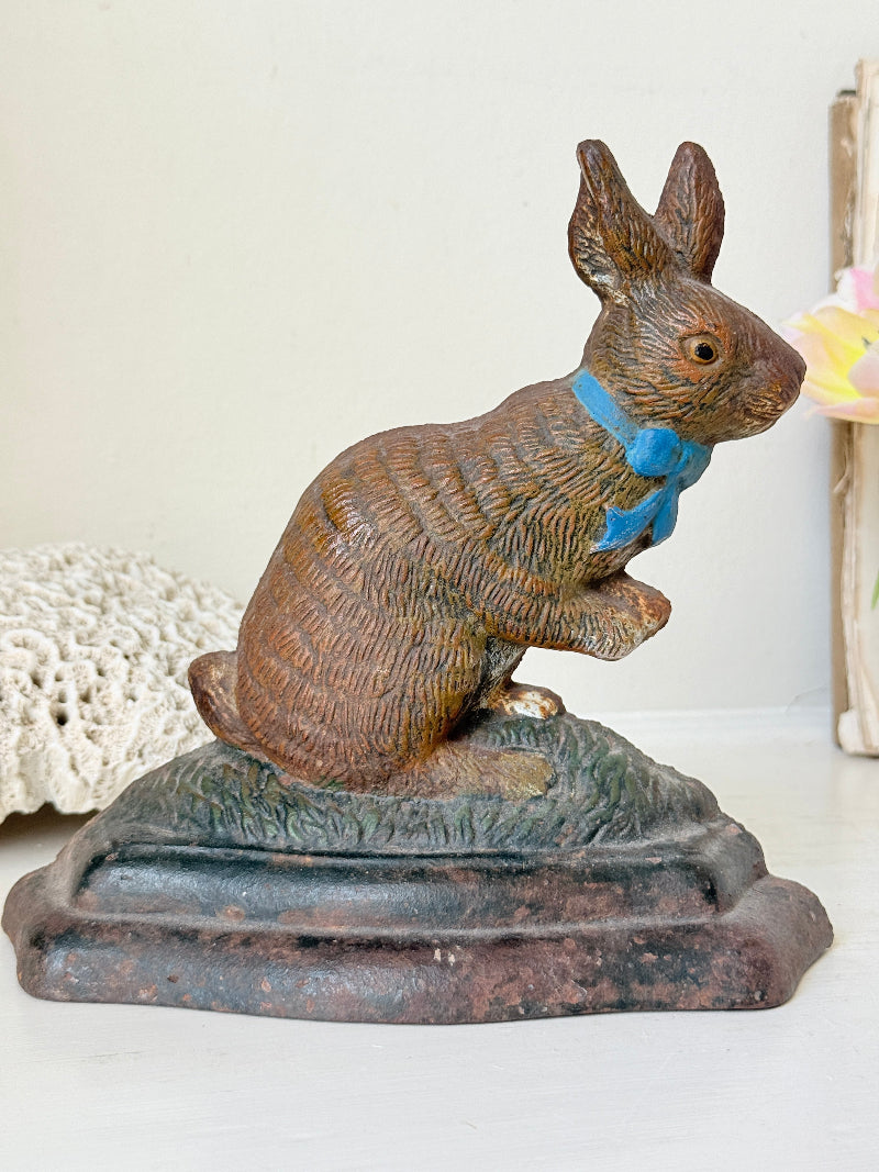 A vintage-style cast iron rabbit figurine wearing a blue bow tie, sitting on a textured grassy base with a dark rectangular pedestal. The rabbit is detailed with brown and beige tones, set against a softly blurred background with a white surface.