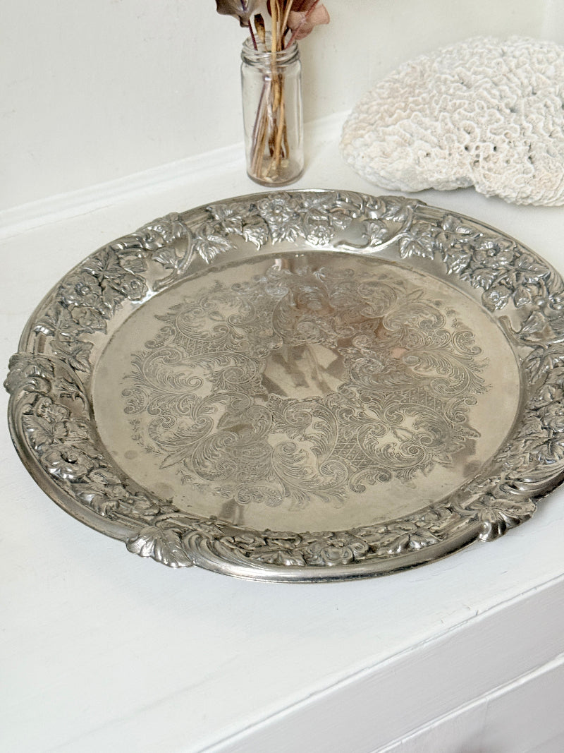 Vintage silver plated round tray with ornate floral border and engraved centre, photographed on a white surface