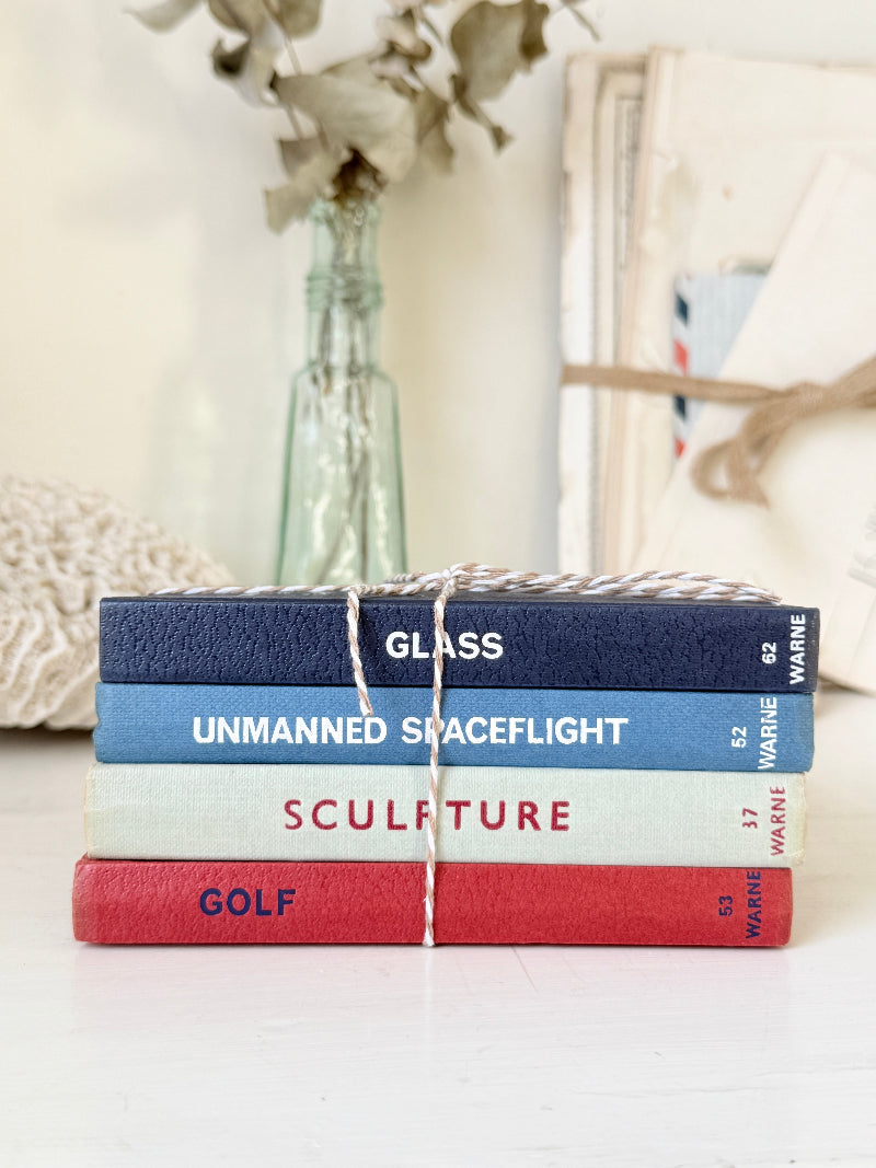 A stack of four hardcover books tied with twine sits on a white surface. The spines read GLASS, UNMANNED SPACEFLIGHT, SCULPTURE, and GOLF, with WARNE visible on the right side of the spines.