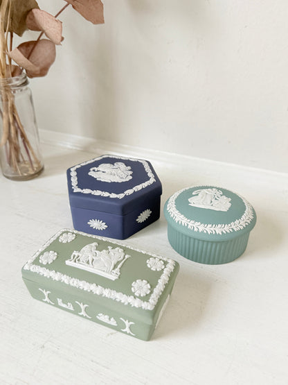 
Trio of vintage Wedgwood Jasperware lidded pots in Portland blue and sage green, featuring neoclassical white relief figures, styled with dried stems on a pale surface.