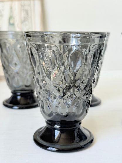 Set of four smoke grey pressed glass footed goblets with a diamond pattern, photographed on a white surface