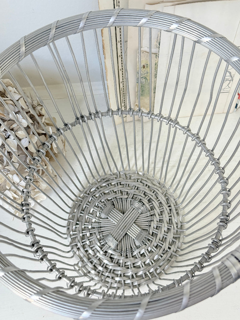 A close-up view of a silver metal wire basket with a woven circular base, photographed indoors against a light neutral background.