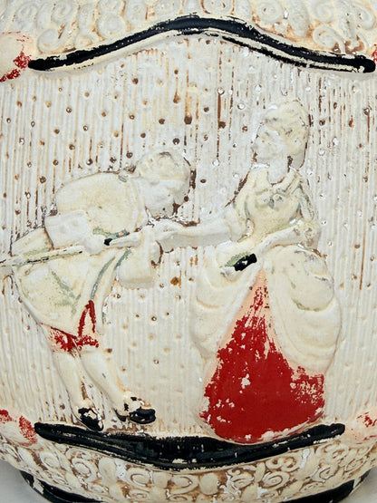 Close-up of a vintage ceramic pitcher or vase featuring a raised relief scene of a couple in a garden, decorated with a textured cream glaze, black outlines, and a prominent red painted area.