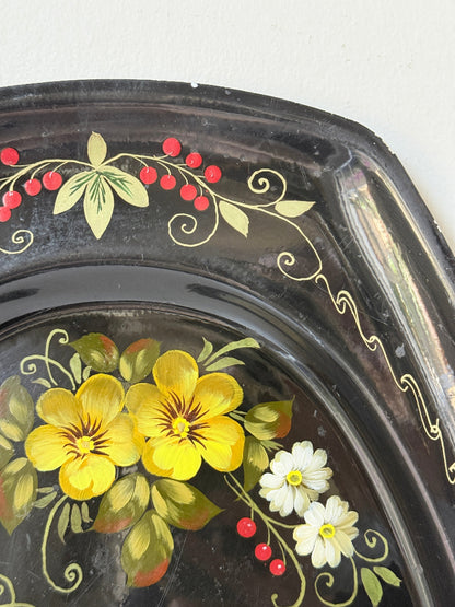 Close-up of a dark decorative tray or plate with hand-painted yellow flowers, white daisies, red berries, and gold vine details on a glossy black surface.