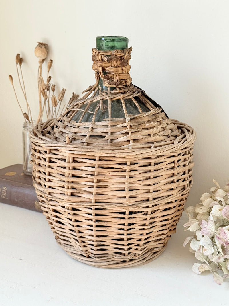 antique French demi john olive oil jar in wicker basket with green glass bottle and painted metal label, displayed on white surface