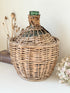 antique French demi john olive oil jar in wicker basket with green glass bottle and painted metal label, displayed on white surface