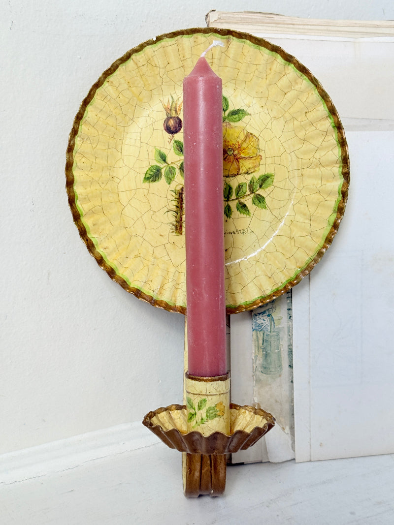A decorative wall-mounted candle holder featuring a tall pink taper candle in a scalloped gold-toned holder, with a large vintage-style yellow plate with botanical illustrations behind it.