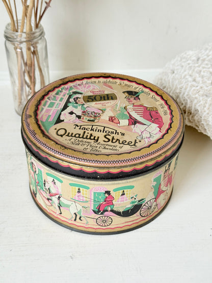 Vintage Quality Street tin from 1986 with colourful illustrated street scenes and pastel design, photographed on a white surface.