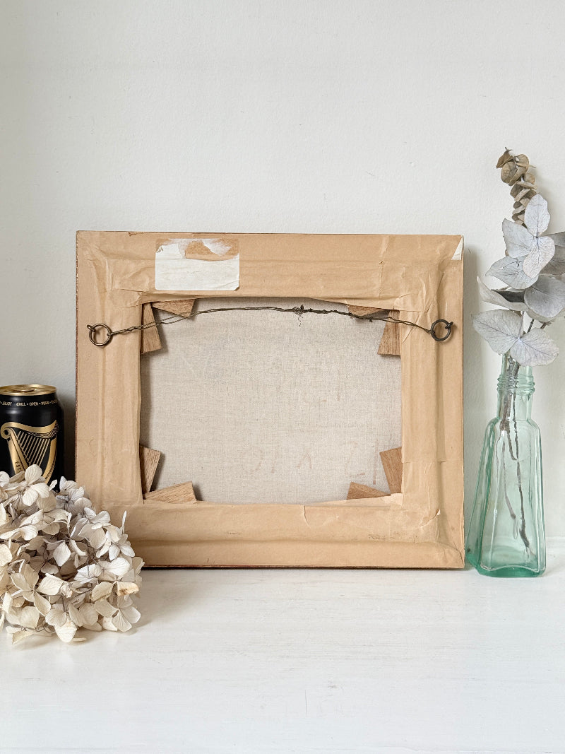 An empty canvas frame with a light brown wood border and wire hanger hardware, displayed on a white surface with dried flowers and a glass vase in the background.
