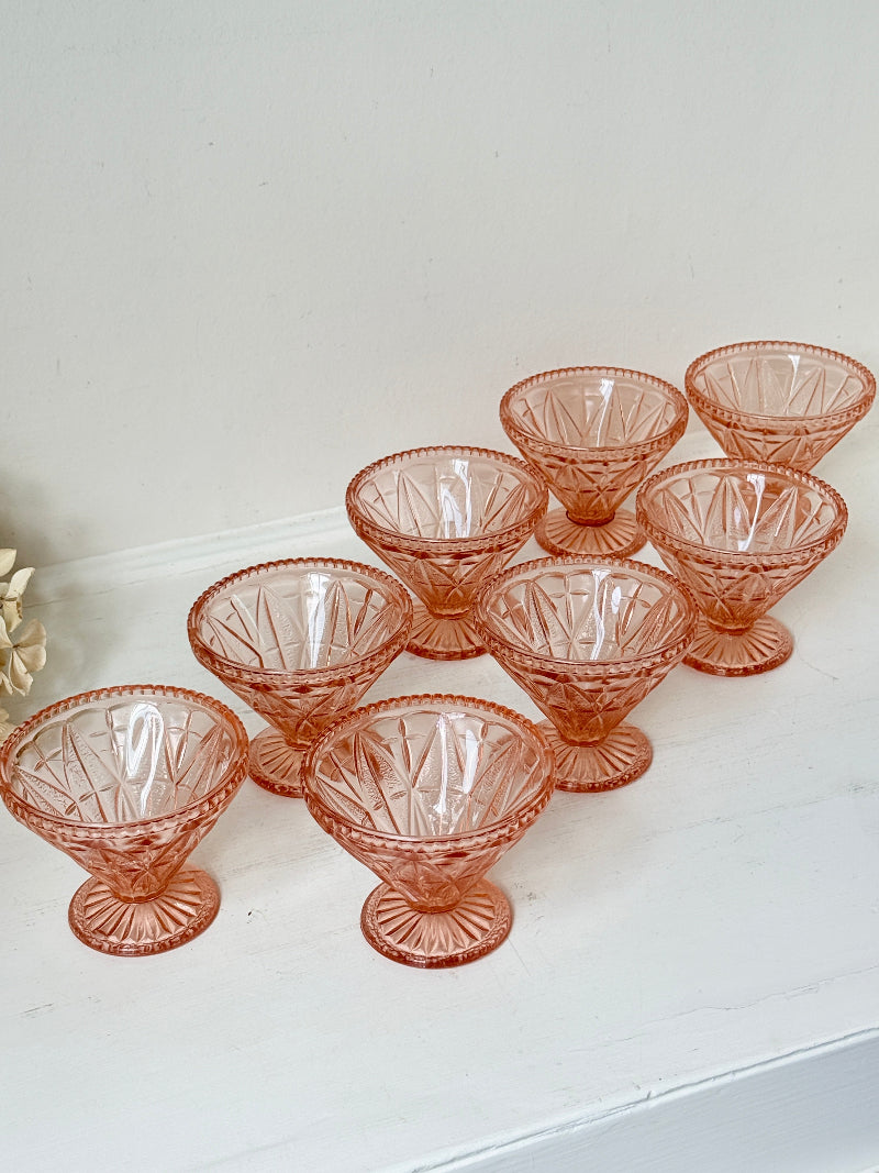 A set of eight vintage pink cut-glass dessert cups arranged on a white shelf against a white wall.
