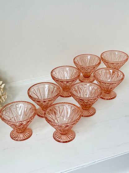 A set of eight vintage pink cut-glass dessert cups arranged on a white shelf against a white wall.