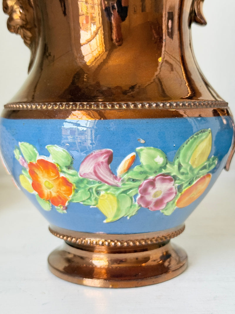 Close-up of a decorative vase with a glossy blue lower body featuring hand-painted flowers and leaves, a reflective metallic copper upper section, and a matching metallic base.