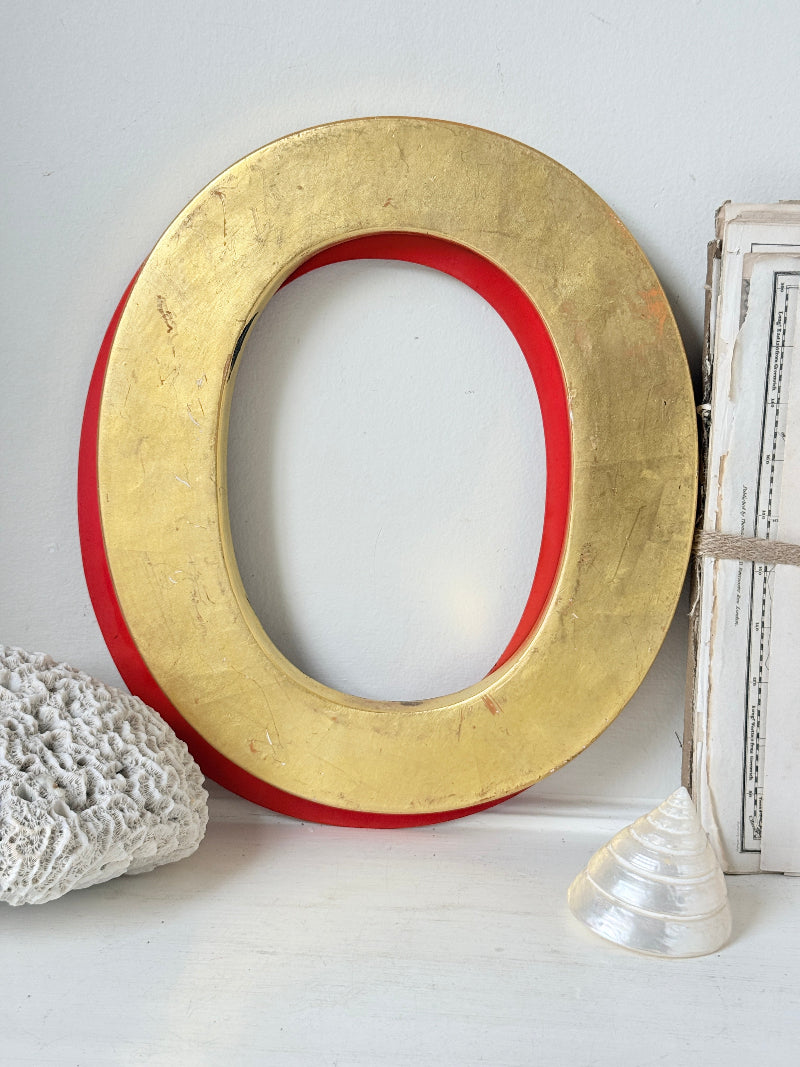 A large decorative letter O wall art piece with a distressed gold finish and a red inner border, styled on a white surface with a white crochet pillow, a seashell, and a vintage book.