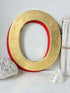 A large decorative letter O wall art piece with a distressed gold finish and a red inner border, styled on a white surface with a white crochet pillow, a seashell, and a vintage book.