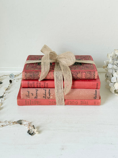 A stack of four vintage-style decorative books in coral red and pink tones, tied with a natural jute ribbon bow, displayed on a white surface against a white wall.