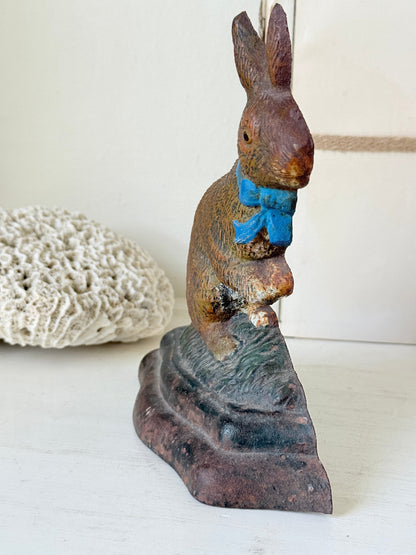 A vintage cast iron Easter bunny figurine wearing a bright blue bow tie, standing on a dark textured base. The figurine is shown on a white surface next to a white coral shell, with a neutral off-white background.
