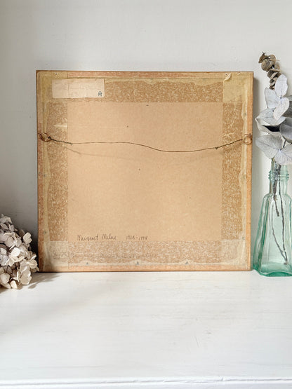 The back of a framed artwork or print, showing a tan paper backing, mounting tape border, and hanging wire, displayed on a white shelf next to a glass vase with dried flowers.