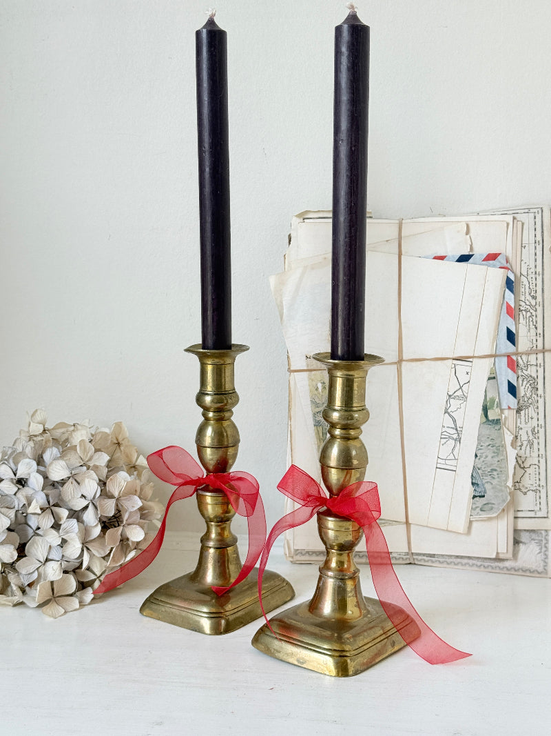 Pair of turned brass candlesticks with square stepped bases and black taper candles on a white surface, styled with dried flowers