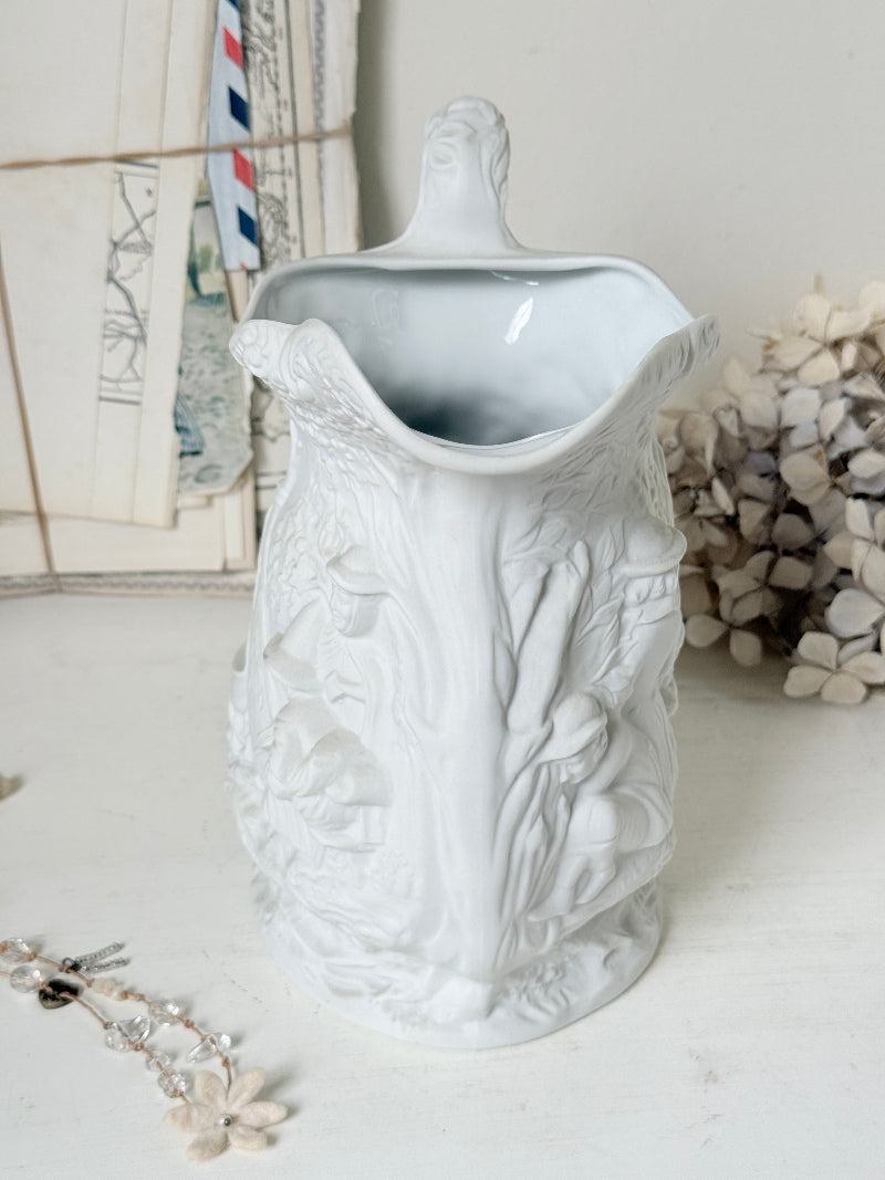 A white ceramic pitcher with intricate embossed floral and leaf patterns, featuring a small handle on the lid and a wide pouring spout, set against a softly styled background with vintage postcards and dried flowers.