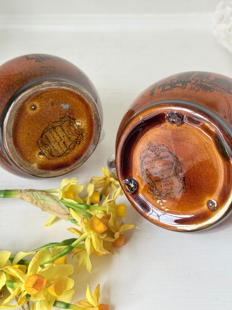 Pair of antique Ridgways stoneware creamer jugs photographed on a white surface in amber treacle glaze with black coaching scenes and silver lustre rims