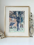 Framed vintage watercolour painting of a large tree and brick house, photographed on a white surface.