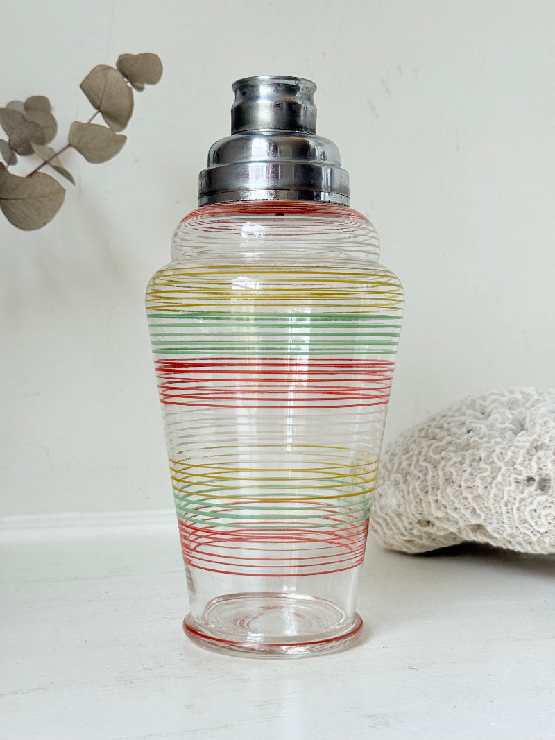 Vintage glass cocktail shaker with multicoloured stripes and silver lid photographed on a white surface.