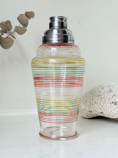 Vintage glass cocktail shaker with multicoloured stripes and silver lid photographed on a white surface.