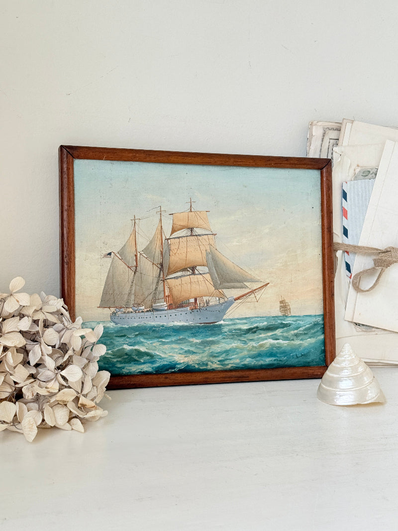 A framed vintage-style painting of a tall sailing ship on blue ocean waves, displayed on a white surface with decorative seashells and a small glass shell beside it.