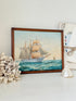 A framed vintage-style painting of a tall sailing ship on blue ocean waves, displayed on a white surface with decorative seashells and a small glass shell beside it.