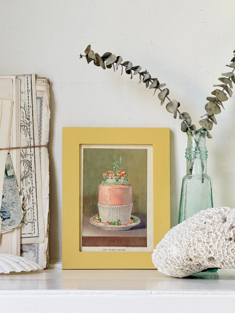 Framed Victorian cookery illustration of a celebration cake from 1896 in a mustard yellow frame displayed on a white surface.